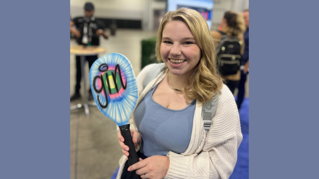 A smiling woman holding a pickleball paddle with a colorful airbrushed design featuring the word "gin." The paddle has a blue background with a vibrant pink, green, and black design. This image highlights the use of airbrushing on suitable materials, showcasing how vibrant and appealing the results can be on appropriate surfaces.